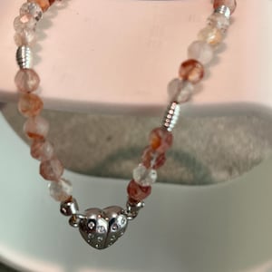Könnte beinhalten: Armband mit orangefarbenen und klaren Perlen und einem silbernen Herzanhänger. Die runden Perlen werden durch silberfarbene Elemente getrennt. Der Herzanhänger ist mit kleinen Punkten verziert. Länge ca. 18 cm.