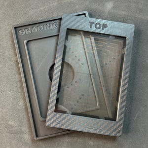 Può includere: Due strumenti di valutazione grigi. Uno reca la scritta "GRADING", l'altro "TOP". Lo strumento a destra ha una finestra trasparente con una griglia e numeri. Gli strumenti hanno un aspetto strutturato simile alla fibra di carbonio.