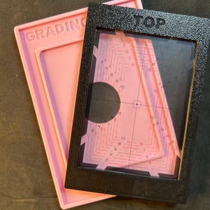 Può includere: Due strumenti di classificazione. Uno è un telaio rettangolare rosa con la scritta "GRADING" in rilievo. L'altro è un telaio rettangolare nero con un centro trasparente e un interno rosa con numeri e linee. La parola "TOP" è stampata sulla parte superiore del telaio nero.