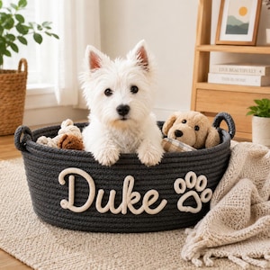 Może przedstawiać: Biały szczeniak West Highland Terrier siedzi w ciemnoszarej, linowej budce dla psa. Na budce widnieje napis "Duke" w kremowym kolorze i odcisk łapy. W budce znajduje się również pluszowy pies i zabawki. Beżowy koc jest zarzucony na bok.