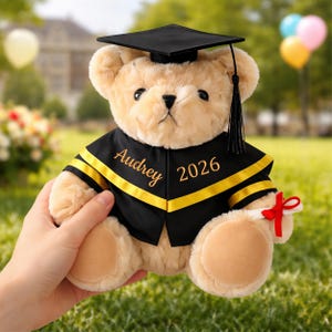 Könnte beinhalten: Ein beigefarbener Teddybär mit schwarzer Abschlusskappe und -robe mit goldenen Verzierungen. Die Robe ist mit dem Namen "Audrey" und dem Jahr "2026" personalisiert. Der Bär hält ein gerolltes Diplom, das mit einem roten Band gebunden ist.