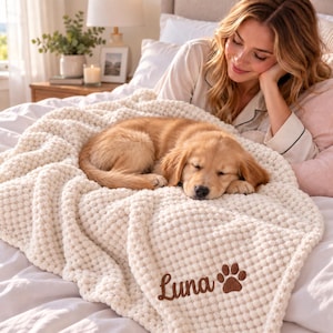 May include: A cream-colored, textured, popcorn-knit throw blanket featuring the embroidered name "Luna" in brown script lettering, accompanied by a brown embroidered paw print icon, draped over a bed with a sleeping golden retriever puppy.