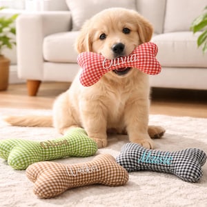 Może przedstawiać: Szczeniak golden retriever trzyma w pysku czerwoną i białą, w kratkę zabawkę w kształcie kości z wyhaftowanym imieniem "Olivia". Na podłodze leżą inne zabawki w kształcie kości w kolorze zielonym, brązowym i czarnym.