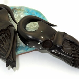 May include: A pair of black wooden ear gauges with intricate carvings and white accents. The gauges are shaped like a skull and have a curved design.