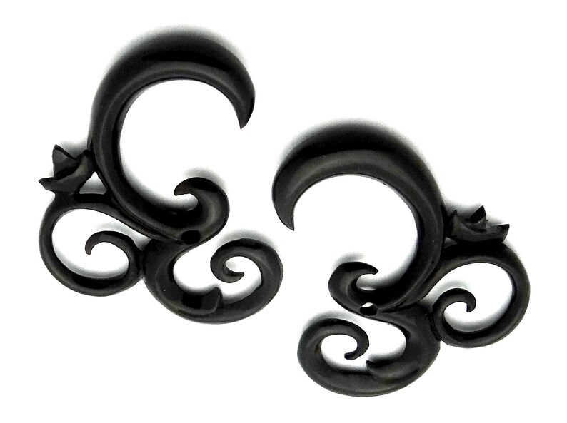May include: A pair of black, ornate, wooden ear gauges. The gauges are shaped like a stylized swirl with a small flower detail.