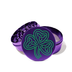 May include: A purple herb grinder with a green Celtic knot shamrock design. The grinder is open, revealing the grinding teeth and mesh screen. The design is on the top lid.