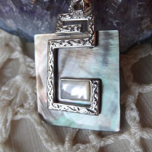 May include: A silver pendant with a square frame and a mother-of-pearl inlay. The frame has a decorative design and the inlay is a light, iridescent white.