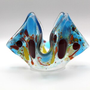 May include: A decorative glass bowl with a unique, folded shape. The bowl features a clear base and is adorned with vibrant splashes of blue, red, yellow, and orange. The colors are distributed throughout the glass, creating a speckled effect.