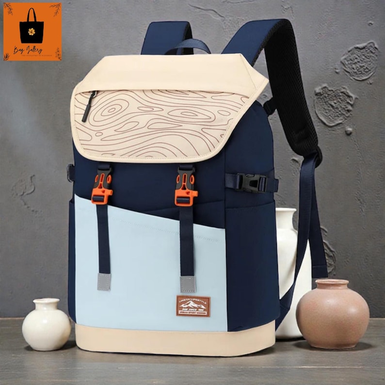 May include: A multi-tone backpack with a flap closure. The backpack features a navy blue body, a light blue front panel, and a beige base and flap. The flap has a wood grain design. Orange buckles and straps secure the flap.