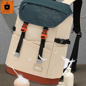 May include: Beige backpack with a dark green top flap featuring a topographic design. The backpack has orange buckles, black straps, and a brown base. A small patch with text is visible on the front.