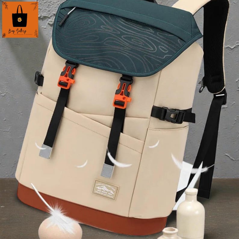 May include: Beige backpack with a dark green top flap featuring a topographic design. The backpack has orange buckles, black straps, and a brown base. A small patch with text is visible on the front.