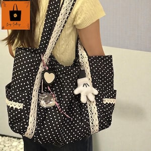 Puede incluir: Bolso tote negro con lunares blancos, con ribete de encaje blanco y correa a juego. El bolso tiene dos bolsillos exteriores y está adornado con un charm en forma de corazón y un peluche en forma de guante blanco. El bolso tiene el texto "Bag Gallery".