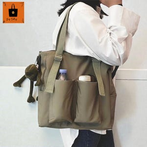 Può includere: Borsa tote verde oliva con più tasche e una lunga tracolla. La borsa ha un peluche a forma di rana attaccato. Una bottiglia d'acqua e un piccolo contenitore sono visibili nelle tasche anteriori.