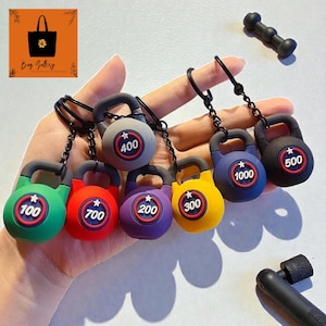 Pode incluir: Uma coleção de chaveiros de kettlebell coloridos em verde, vermelho, roxo, amarelo, azul, cinza e preto. Cada chaveiro exibe um peso em quilogramas, variando de 100 a 1000. Os chaveiros são presos a correntes de metal preto.
