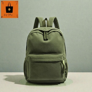 Puede incluir: Mochila de pana verde oliva con un bolsillo frontal y correas ajustables. La mochila tiene un asa superior y un cierre de cremallera. La bolsa tiene un pequeño detalle trenzado decorativo en la parte delantera.