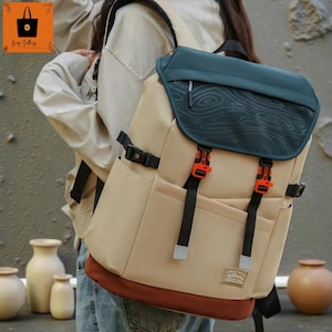 Op de afbeelding: Een beige rugzak met een teal klep met een houtnerfpatroon. De rugzak heeft zwarte banden, oranje gespen en een bruine basis. Hij heeft twee zijzakken en een klein label.