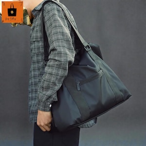 May include: A black shoulder bag with a zippered front pocket and a wide shoulder strap. The bag is made of a smooth, durable material and is designed for carrying items. The bag is being carried over the shoulder.