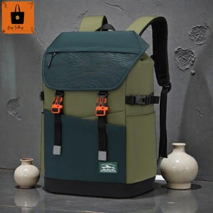 May include: A multi-tone backpack in olive green, teal, and black. The flap features a topographic design. Orange buckles secure the front pockets. The bag has adjustable straps and a top handle. A small label is visible on the front.