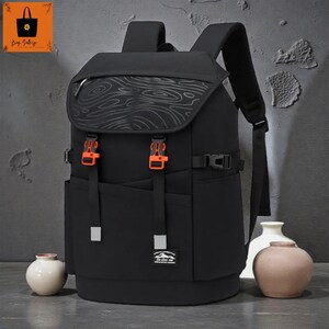 May include: Black backpack with a flap closure and orange buckles. The flap has a wood grain pattern. The backpack has adjustable straps and a front pocket with a small patch. The bag has a modern design.