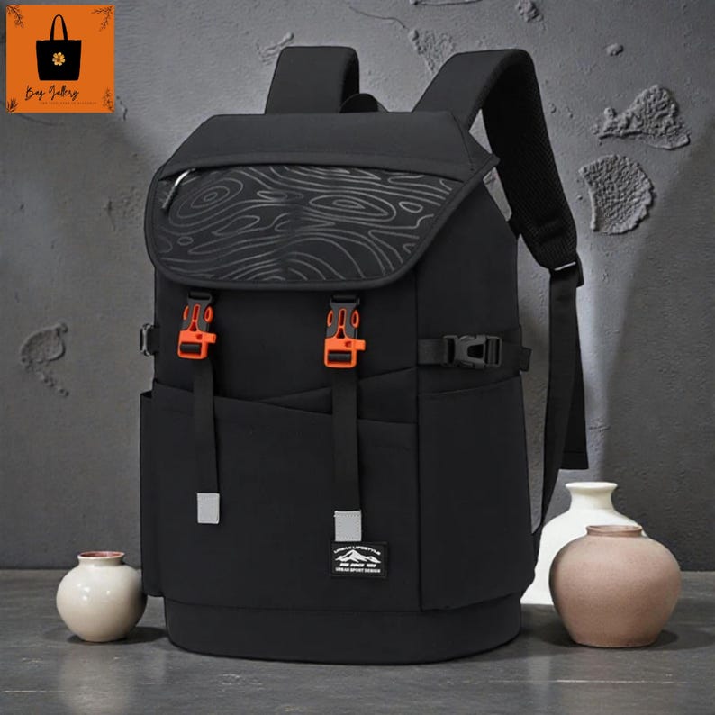 May include: Black backpack with a flap closure and orange buckles. The flap has a wood grain pattern. The backpack has adjustable straps and a front pocket with a small patch. The bag has a modern design.