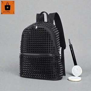 Può includere: Zaino nero ricoperto di borchie nere. Lo zaino ha uno scomparto principale con cerniera e una tasca frontale più piccola. Gli spallacci sono neri e regolabili. Una penna nera e due oggetti circolari bianchi sono accanto allo zaino.