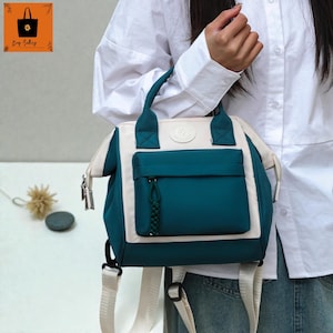 Puede incluir: Una mochila de color verde azulado y crema con un bolsillo frontal y un asa superior. La bolsa tiene correas ajustables y cierre de cremallera. El logotipo "By Jaling" está en la esquina superior izquierda.