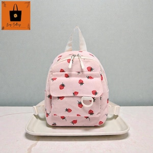 May include: Pink corduroy backpack with a strawberry pattern. The backpack has a small front pocket, white zippers, and adjustable straps. The bag has a top handle and a D-ring. The bag is sitting on a white plate.