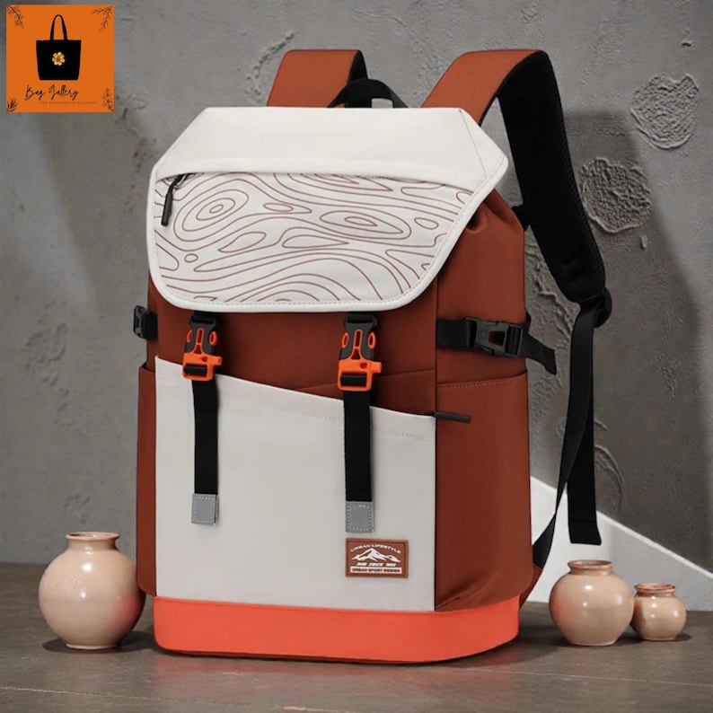 May include: A brown and white backpack with orange detailing. The backpack has a white flap with a brown topographic design, orange buckles, and a small patch with text. It features black straps and a white front panel.
