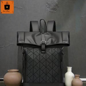May include: Black geometric-patterned backpack with a roll-top closure secured by a silver buckle. The bag has a padded top and shoulder straps. The background is a dark gray wall, and there are decorative vases in the foreground.