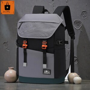 May include: A multi-tone backpack with a grey top flap featuring a topographic design. The backpack has black, grey, and teal panels, with orange buckles and straps. A small logo is visible on the front.