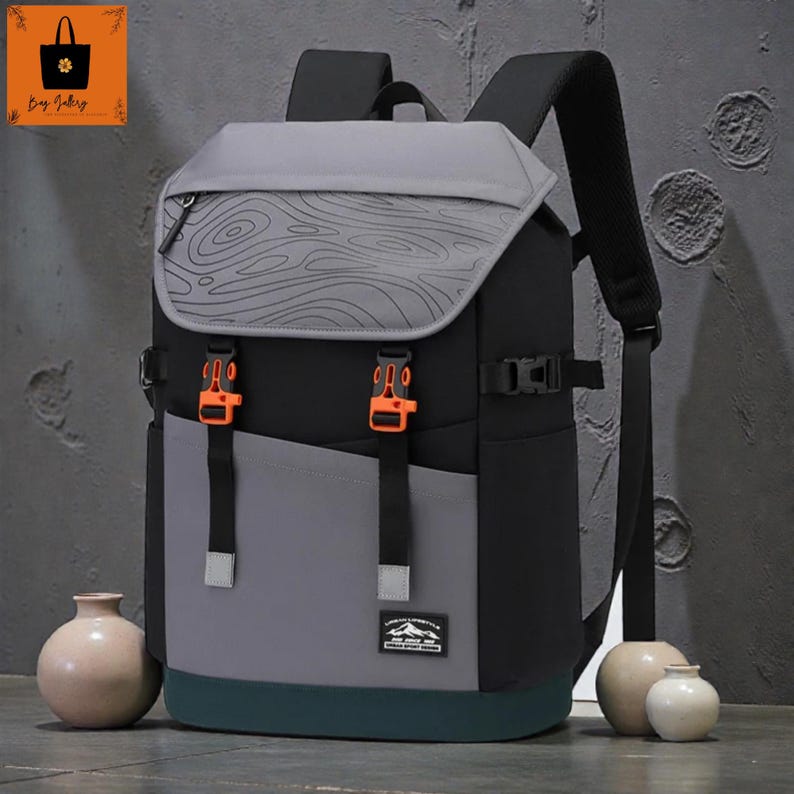 May include: A multi-tone backpack with a grey top flap featuring a topographic design. The backpack has black, grey, and teal panels, with orange buckles and straps. A small logo is visible on the front.
