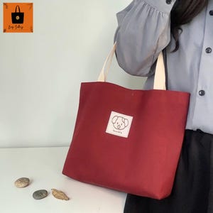 May include: A red tote bag with cream-colored handles and a small square patch featuring a dog illustration. The bag is made of a sturdy fabric and is designed for carrying items. The bag has the text "Spunky Style" on the patch.
