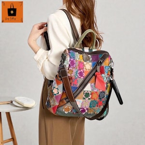 Puede incluir: Una mochila de cuero de retazos multicolor con un diseño floral. La bolsa presenta una variedad de colores, incluyendo rojo, azul, verde y morado, y tiene correas de cuero marrón y un cierre de cremallera. La bolsa tiene un asa superior y una correa para el hombro.