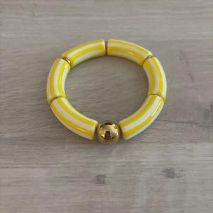 May include: A stretch bracelet composed of cylindrical beads featuring alternating yellow and white vertical stripes, interspersed with a single polished gold-toned spherical bead. The beads are arranged in a circular pattern to form the bracelet.