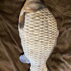 K&ouml;nnte beinhalten: Ein pl&uuml;schiges Fischkissen in Beige-, Braun- und Blaut&ouml;nen. Das Kissen hat detaillierte Schuppen, Flossen und einen Schwanz. Das Fischkissen liegt auf einer braunen Oberfl&auml;che.