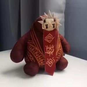 May include: A plush toy in shades of burgundy and maroon, featuring a stylized face with a sun-like crown. The toy has a decorative scarf with geometric patterns and text. The toy is sitting on a white surface.