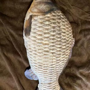 K&ouml;nnte beinhalten: Ein kissenf&ouml;rmiger Fisch mit einem detaillierten Schuppenmuster in Beige- und Graut&ouml;nen. Das Kissen hat ein realistisches Design, einschlie&szlig;lich Flossen und einem Schwanz mit blauen Akzenten. Das Fischkissen liegt auf einer braunen Oberfl&auml;che.