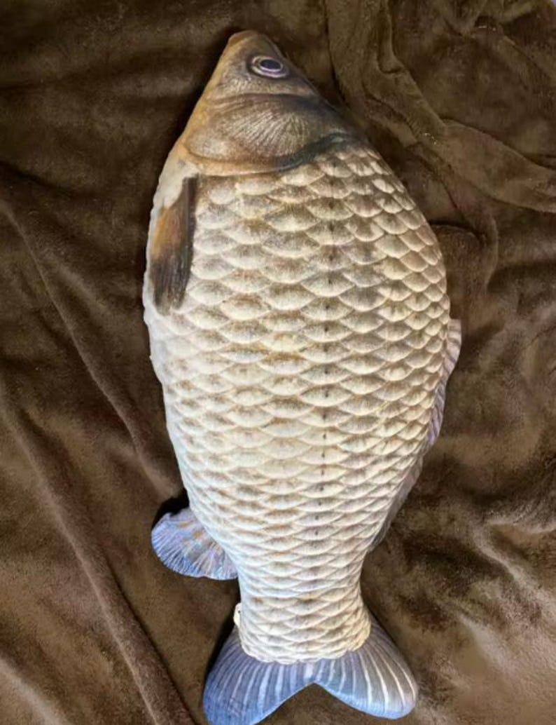 K&ouml;nnte beinhalten: Ein kissenf&ouml;rmiger Fisch mit einem detaillierten Schuppenmuster in Beige- und Graut&ouml;nen. Das Kissen hat ein realistisches Design, einschlie&szlig;lich Flossen und einem Schwanz mit blauen Akzenten. Das Fischkissen liegt auf einer braunen Oberfl&auml;che.