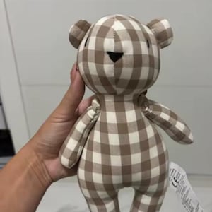 Puede incluir: Un oso de peluche con un patrón de cuadros marrones y blancos. El oso tiene orejas pequeñas, una nariz cosida negra y una expresión sencilla. El juguete es suave y de felpa, adecuado para niños.