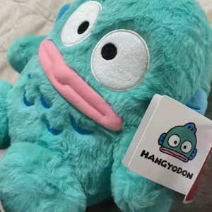 May include: A plush turquoise Hangyodon character with large white eyes, a pink tongue, and blue fins. The plush toy has a tag with the Hangyodon logo and text. The toy is on a soft, light-colored surface.