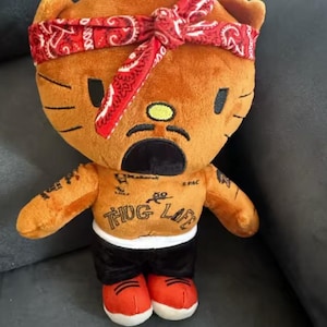May include: A brown plush cat toy with a red bandana, black shorts, and red shoes. The cat has tattoos on its arms and the words "THUG LIFE" printed on its chest. The toy has black eyes and a sad expression.
