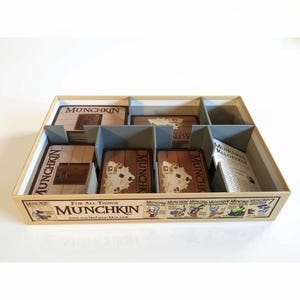 May include: A rectangular cardboard game box organizer with grey dividers containing stacks of Munchkin playing cards. The front of the box features the text For All Things Munchkin, the website address, and various small cartoon character illustrations.