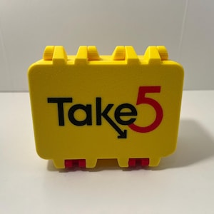 May include: A rectangular yellow plastic storage case with a hinged lid and a textured surface. The front features the black text "Take" followed by a black arrow pointing down and to the right, and a large red number "5" on the right side.