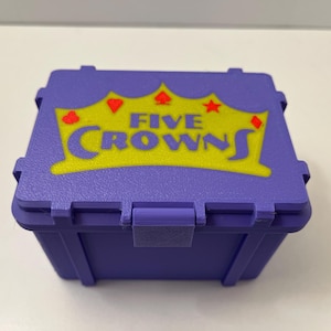 May include: A purple plastic box featuring the text "FIVE CROWNS" in yellow, with a crown, heart, diamond, spade, and star graphic. The box has a hinged lid and a latch. The box is designed for storing game pieces.