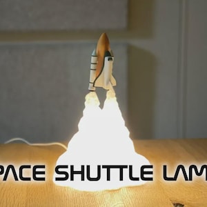 May include: A space shuttle lamp with a white and brown design, emitting a bright light from its base. The lamp is on a wooden surface, with the words "SPACE SHUTTLE LAMP" in white text at the bottom.