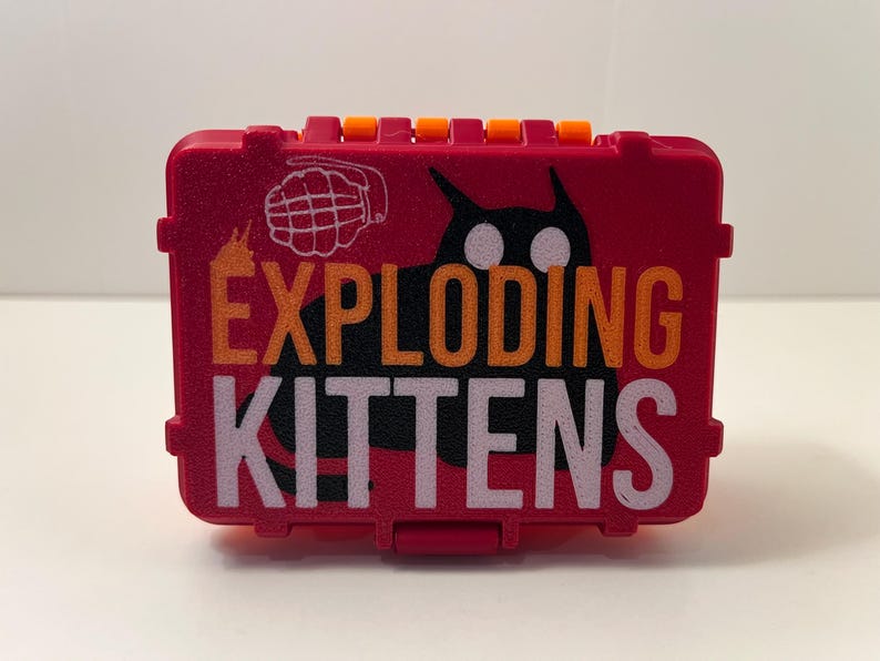 K&ouml;nnte beinhalten: Eine rote, rechteckige Exploding Kittens-Spielbox. Die Box zeigt das Logo des Spiels in Wei&szlig;, Orange und Schwarz mit einer Cartoon-Katzen-Silhouette. Die Oberseite hat orangefarbene Akzente.