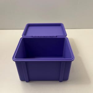 May include: A purple storage box with an open lid. The box is rectangular with a hinged lid and vertical supports on the sides. The box is made of a solid, smooth material. The box is approximately 20 cm long.