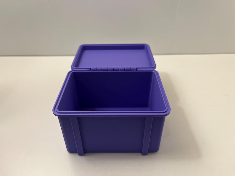 May include: A purple storage box with an open lid. The box is rectangular with a hinged lid and vertical supports on the sides. The box is made of a solid, smooth material. The box is approximately 20 cm long.