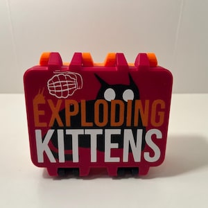 May include: A rectangular, magenta-colored plastic storage case with orange hinges. The front features a black cat silhouette with white eyes, a white grenade icon, and the text EXPLODING KITTENS in large, bold, orange and white capital letters.