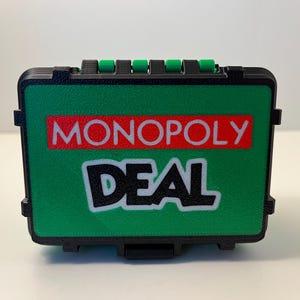 Może przedstawiać: Zielona i czarna skrzynka do gry w karty Monopoly Deal. Skrzynka ma teksturowaną powierzchnię i zawiera napisy "MONOPOLY DEAL" w białych i czerwonych literach. Skrzynka ma czarny uchwyt z zielonymi akcentami.